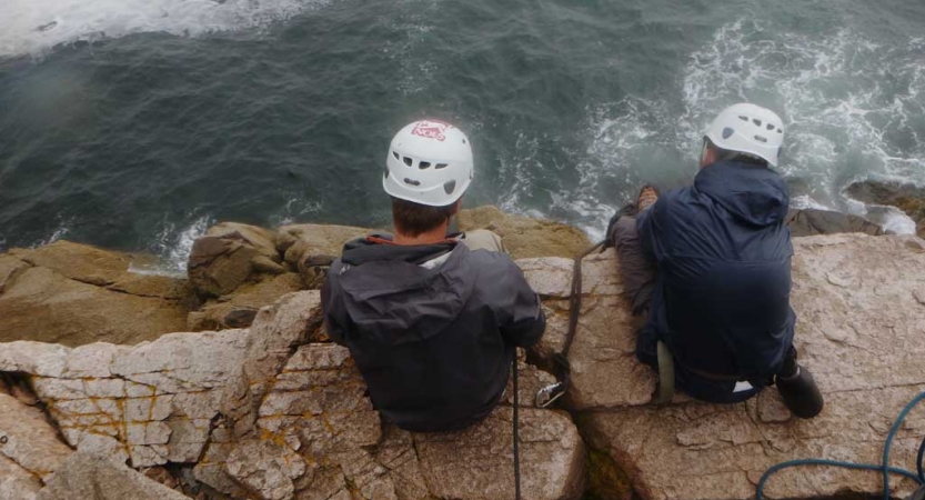 Two people wearing helmets sit on rocks near the shore.